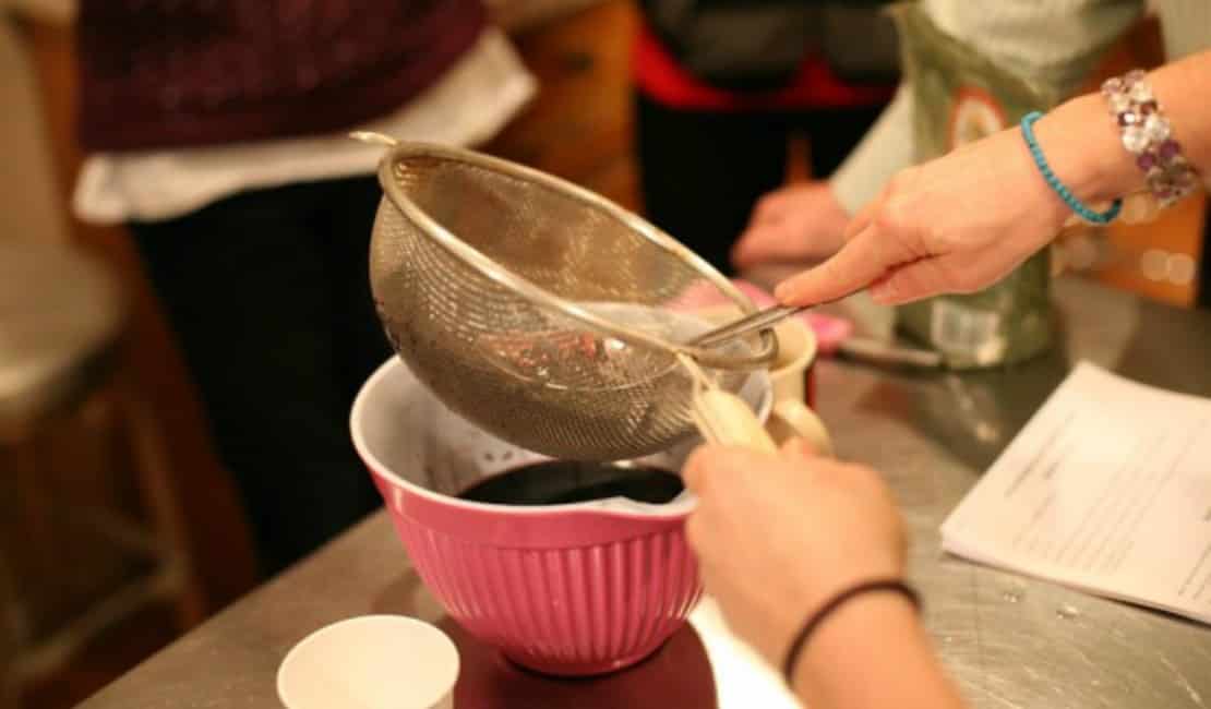 Close-up of hands using a sieve over a pink mixing bowl in a kitchen setting. As one person holds the sieve, another supports the bowl, possibly preparing natural cold and flu remedies. Recipe papers and a measuring cup sit on the countertop, while blurred figures add to the bustling kitchen scene.