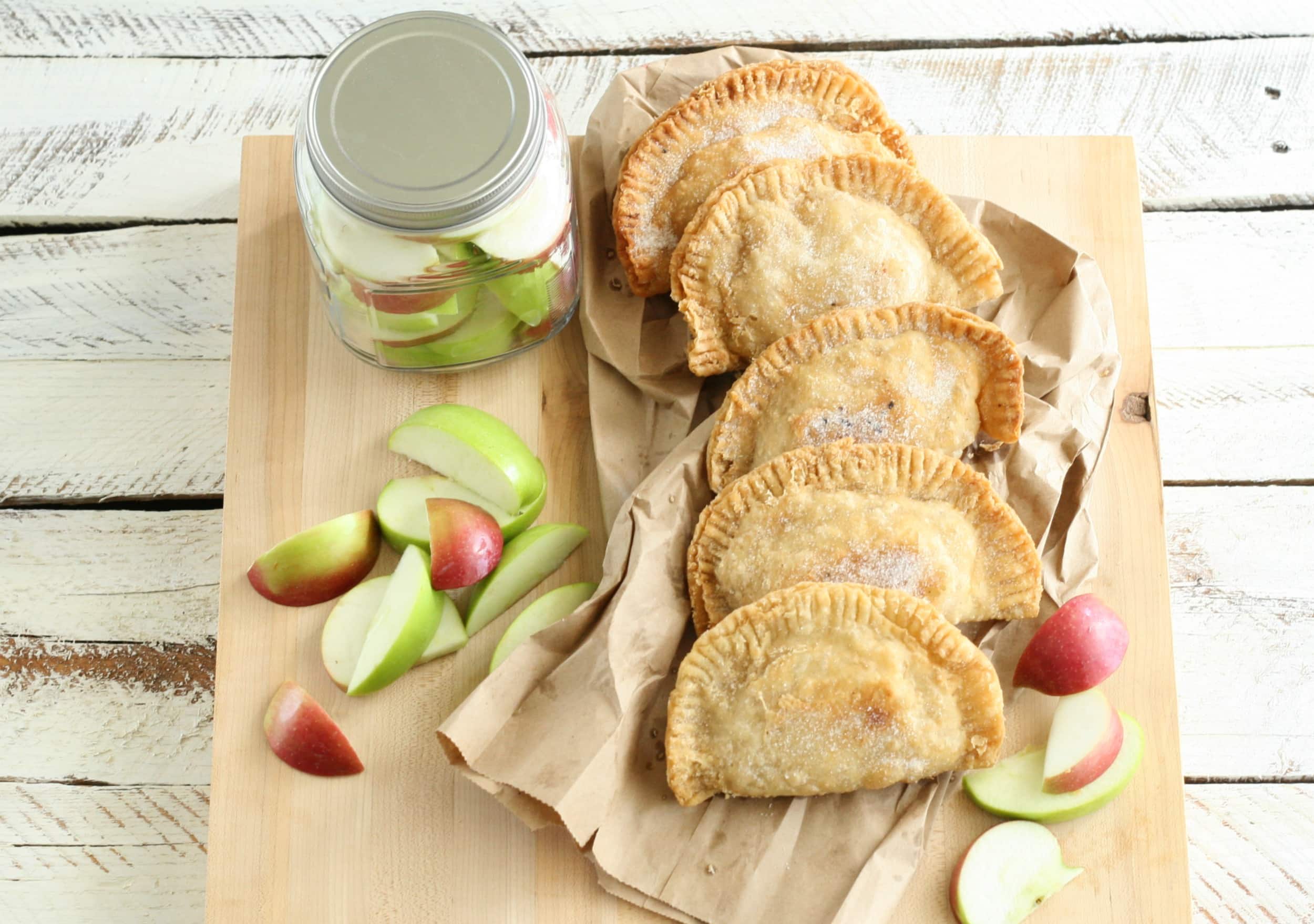 A wooden board showcases five baked empanadas on brown paper, surrounded by a jar filled with apple slices. Additional slices are artfully scattered across the board, offering a rustic touch to this delightful setting, perfect for apple recipes on a charming white wooden surface.
