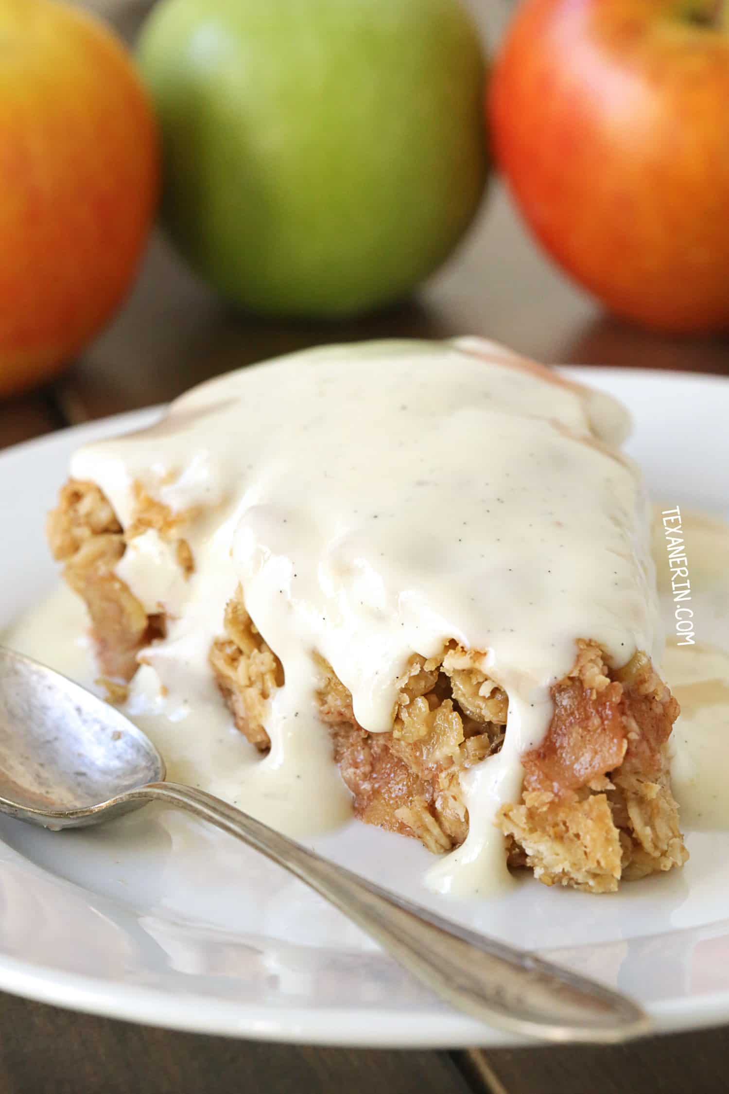 A slice of apple crumble from cherished apple recipes, topped with creamy vanilla sauce, rests on a white plate beside a silver spoon. In the background, vibrant red and green apples add a touch of color.