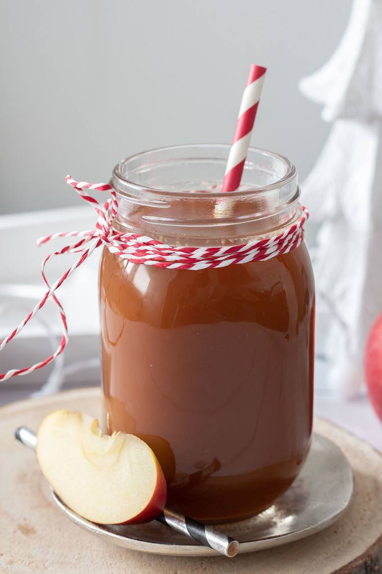 A mason jar filled with apple cider, decorated with red and white twine and a striped straw, sits on a saucer beside a slice of apple. Perfect for showcasing your favorite apple recipes, the blurred background hints at holiday decor.