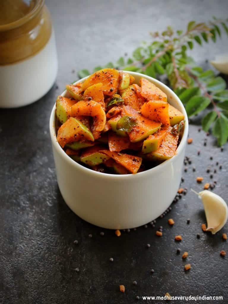 A ceramic bowl filled with Indian mango pickle, garnished with spices, evokes tantalizing aromas. Fresh curry leaves, garlic cloves, and a jar linger on the dark surface, enhancing the vibrant and spicy allure of this enticing creation reminiscent of beloved apple recipes.