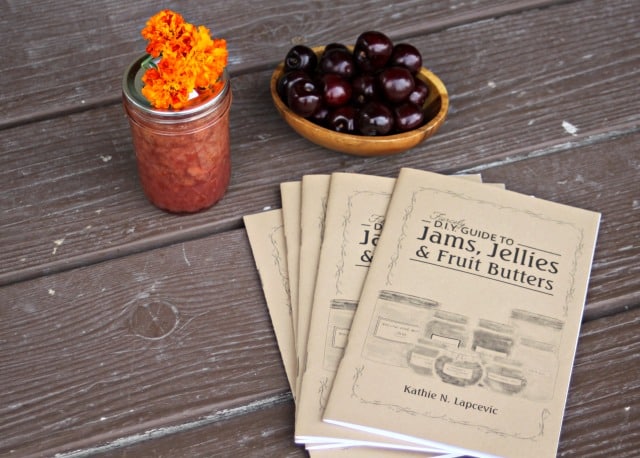 A tabletop with three copies of "DIY Guide to Jams, Jellies & Fruit Butters," a jar of orange marigolds, and a small wooden bowl filled with dark cherries. The surface is wooden, with a rustic appearance.