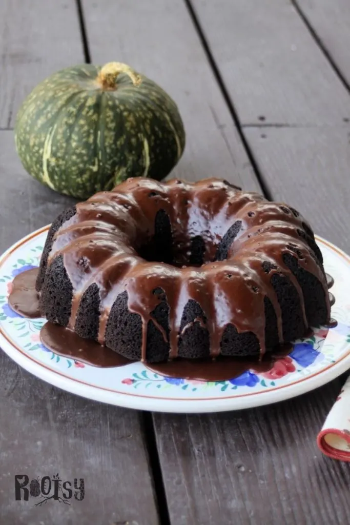 A glazed chocolate pumpkin cake on a floral cake plate with green pumpkin sitting behind it.
