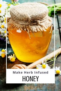 A cloth covered jar of honey sits on a table with a honey dipper and chamomile flowers. Text overlay reads: Make Herb Infused Honey.