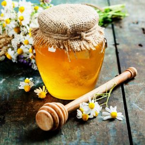 A jar of honey with honey dipper surrounded by fresh herbs and flowers.