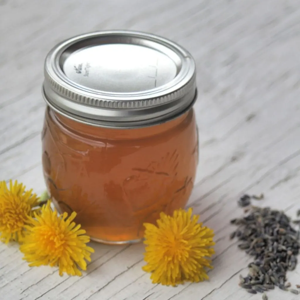 A sealed canning jar full of yellow jelly surrounded by dandelion flowers and lavender buds.