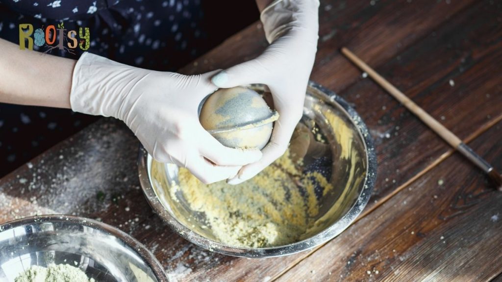 Latex glove covered hands pressing together a bath bomb mold over a metal bowl.