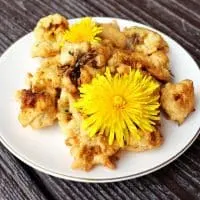 A plate stacked with cooked fritters with 2 dandelion flowers on top.