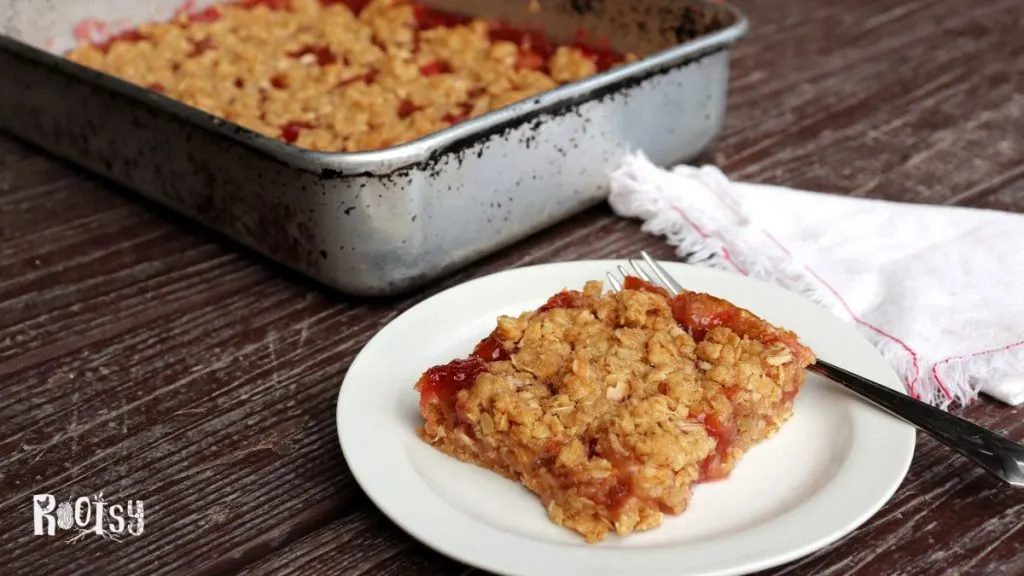 A rhubarb oatmeal bar on a white plate with a fork and napkin with remaining pan full in the background.