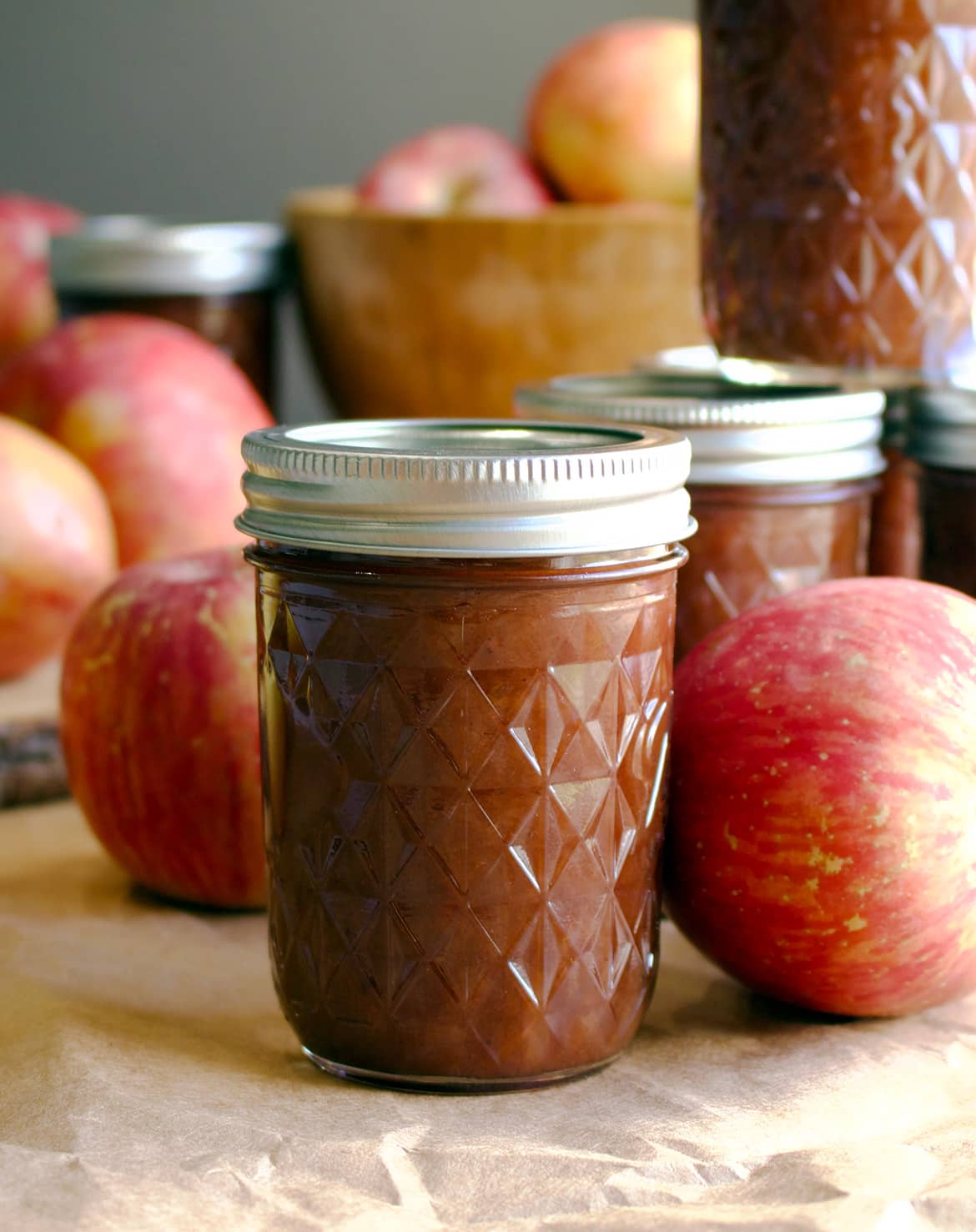 Jars of apple butter sit on a table surrounded by fresh red apples, reflecting classic apple canning recipes. The jars feature a decorative diamond pattern and metallic lids. In the background, a wooden bowl brims with apples, evoking a homemade or rustic setting.