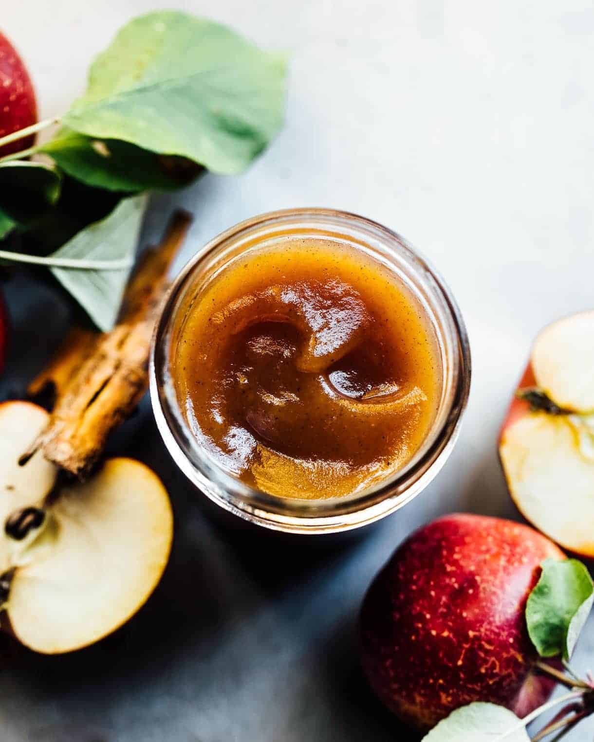 A jar of apple butter, masterfully crafted from classic apple canning recipes, is surrounded by fresh apples, cinnamon sticks, and green leaves. The rich texture and warm brown color invite you to enjoy the juicy red apples that reveal their sweet flesh inside.