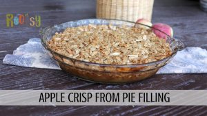 An apple crisp sits on a leaf patterned cloth with fresh apples and a basket in the background. A text overlay reads: apple crisp from pie filling.