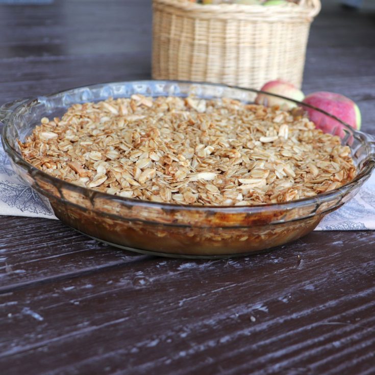 An apple crisp in a clear pie plate sits on a cloth with fresh apples and a basket in the background.
