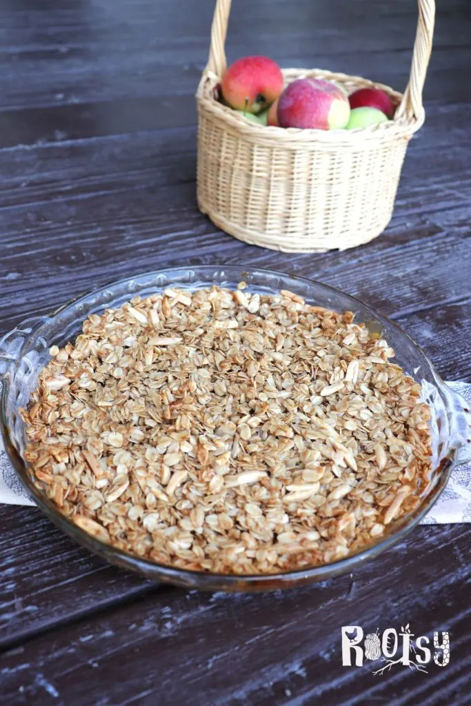 An apple crisp as seen from above in a glass pie plate. A basket of fresh apples sits behind it.