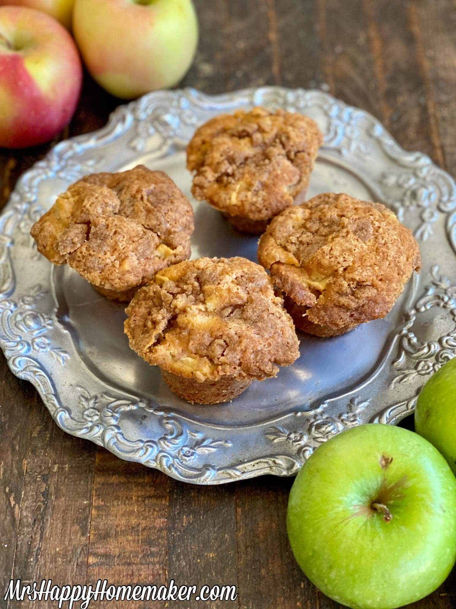 Four freshly baked apple muffins are on a decorative silver plate, surrounded by a mix of red and green apples on a rustic wooden table.