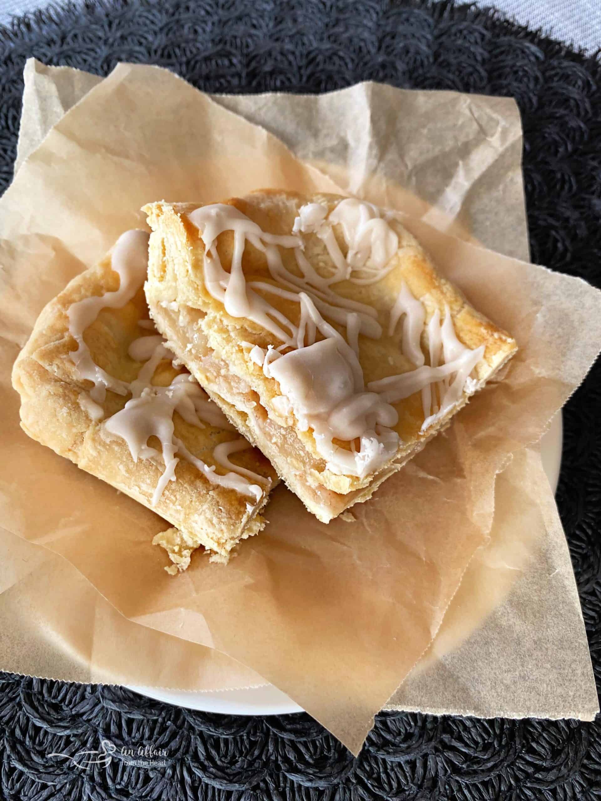 Two slices of dessert bars topped with a white icing drizzle are placed on a piece of parchment paper on a white plate. The dessert has a flaky, golden-brown crust and is set on a dark woven surface.