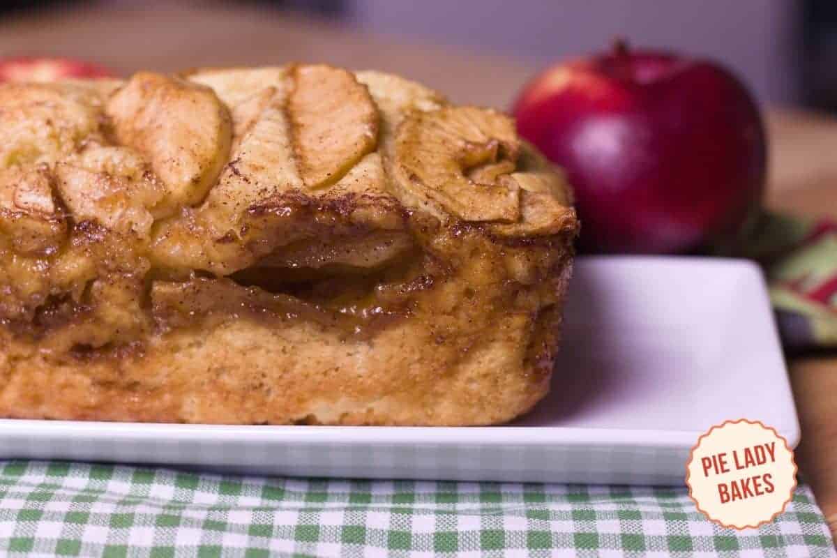 Close-up of a homemade apple cinnamon bread on a white rectangular plate, topped with apple slices. A red apple in the background adds a touch of color. A logo reading "Pie Lady Bakes" is in the bottom right corner. A plaid cloth is partially visible.