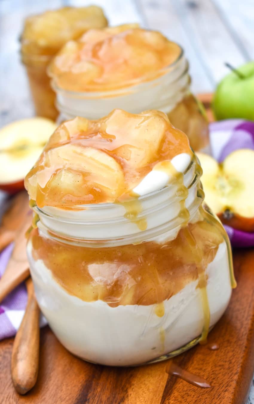 Jars filled with yogurt layered with apple pie filling, topped with caramel sauce, placed on a wooden board. Sliced apples and a wooden spoon are visible in the background.