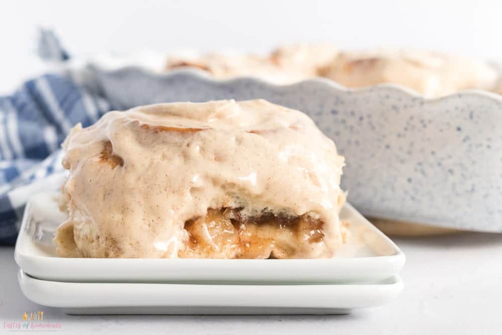 A close-up of a cinnamon roll with creamy icing on a white plate. A bite is taken out, revealing a gooey inside. In the background, a blue speckled baking dish holds more cinnamon rolls. A blue and white striped cloth is partially visible.