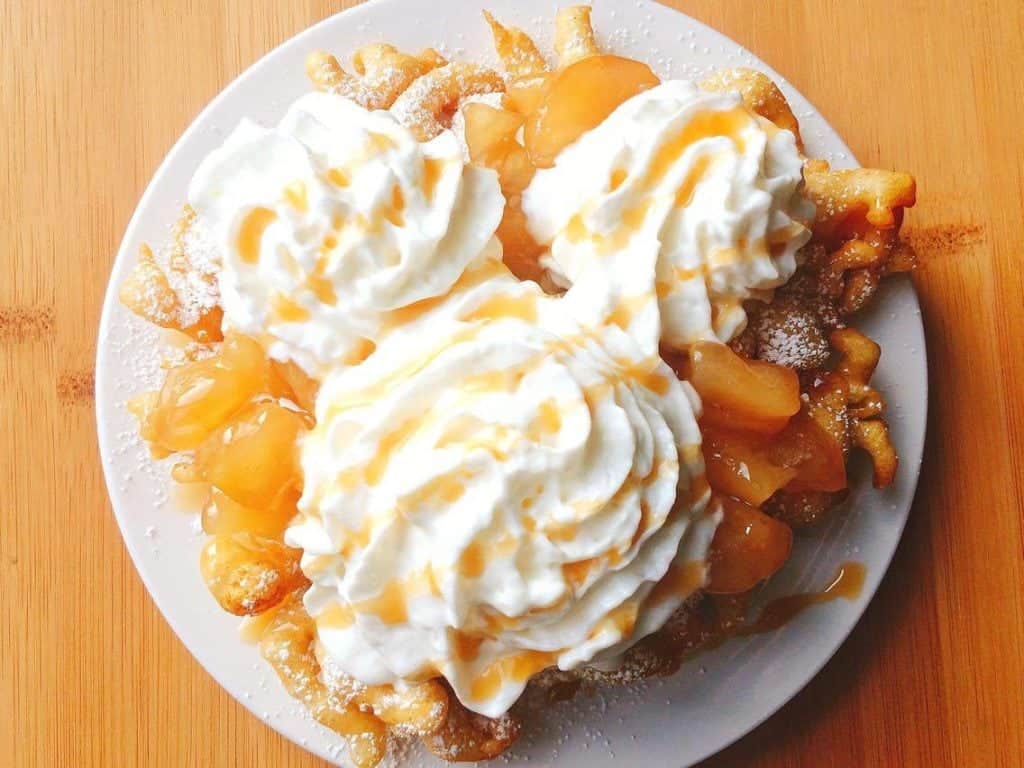 A funnel cake topped with whipped cream, caramel drizzle, and apple pie topping, resembling a Mickey Mouse shape, served on a white plate on a wooden table.