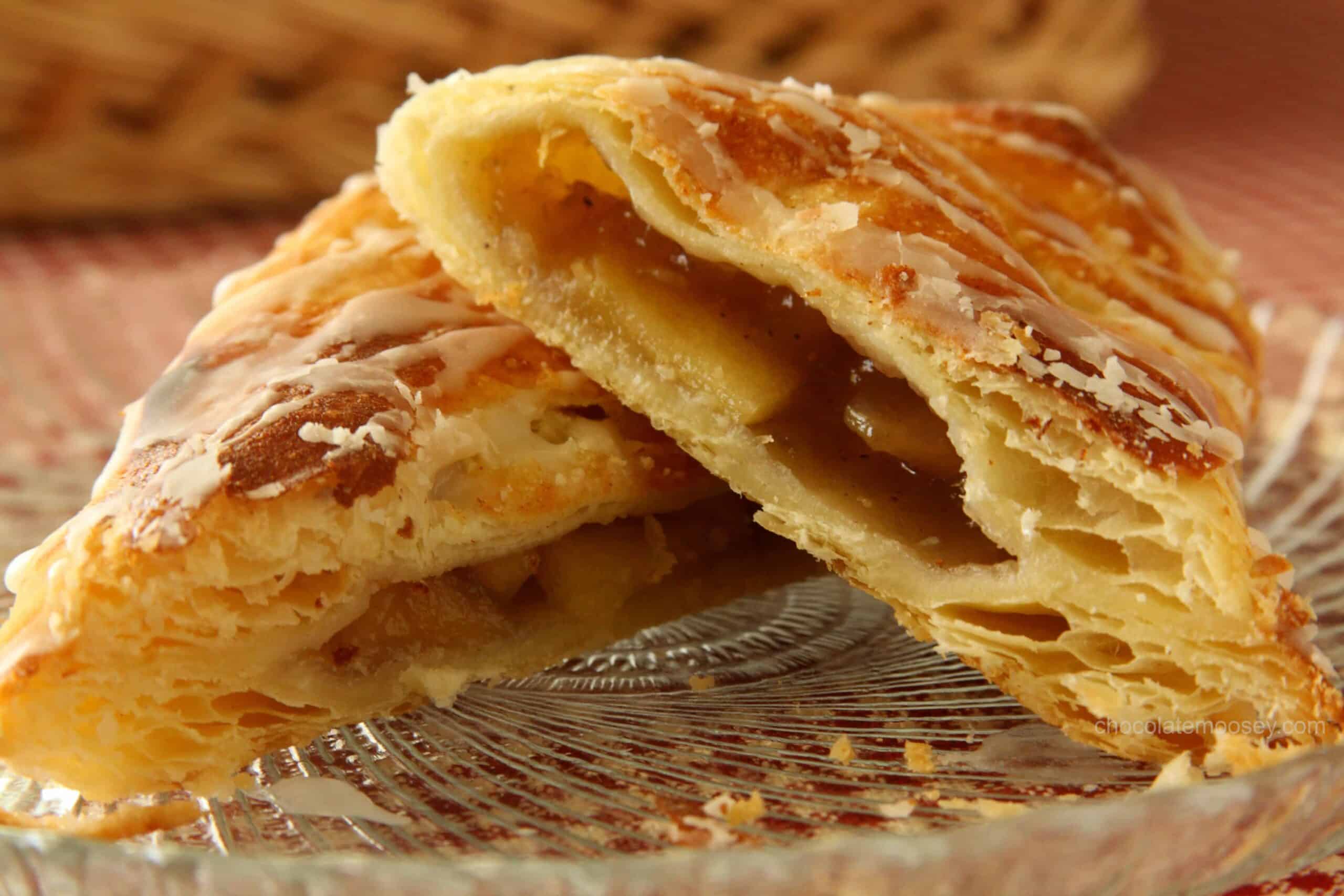 Close-up of two flaky apple turnovers on a glass plate. They are golden brown with a light drizzle of icing on top, revealing a sweet apple filling inside. A woven basket is blurred in the background.