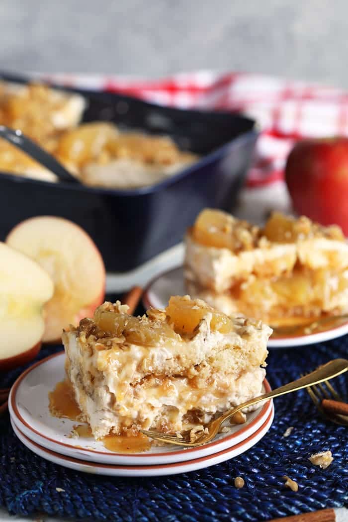 A slice of layered dessert with caramelized apple pieces and walnuts sits on a small plate, alongside a whole apple and a baking dish in the background. A spoon rests beside the dessert on a table mat.