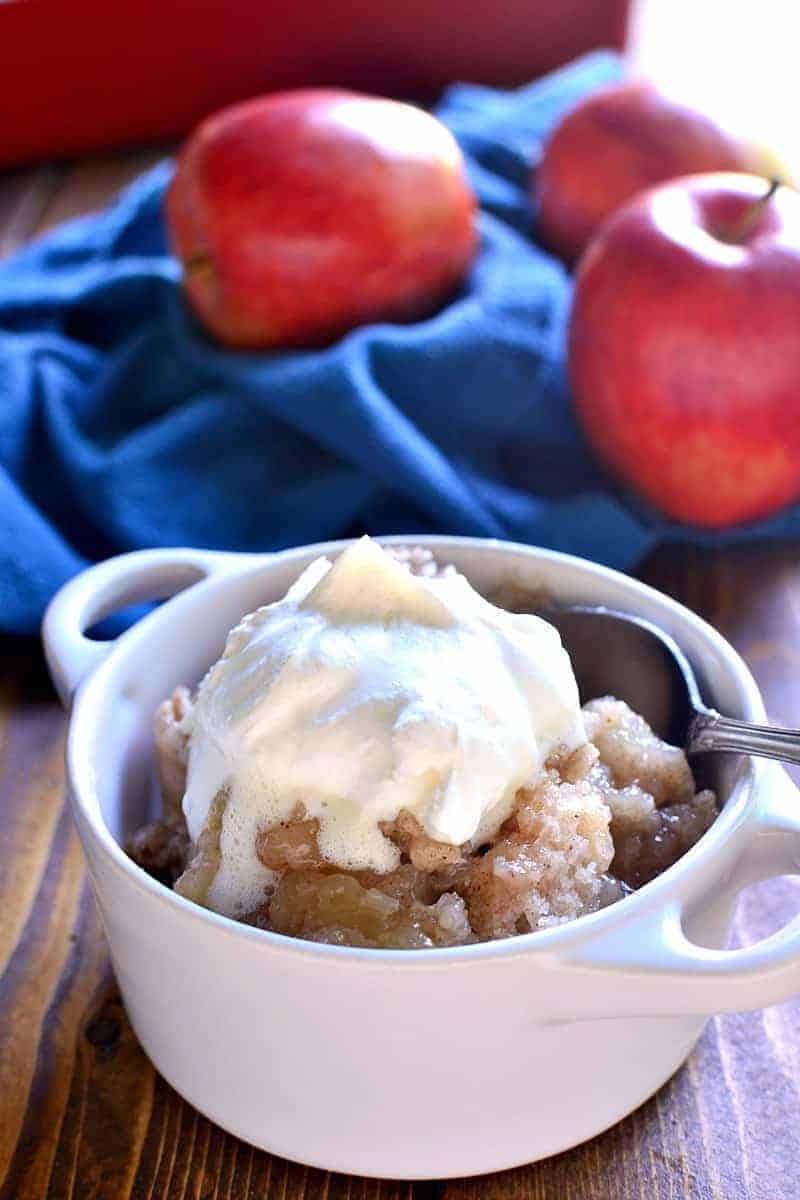 A white bowl of apple crisp topped with whipped cream sits on a wooden table. A spoon rests in the bowl. In the background, several red apples and a folded blue cloth are visible.