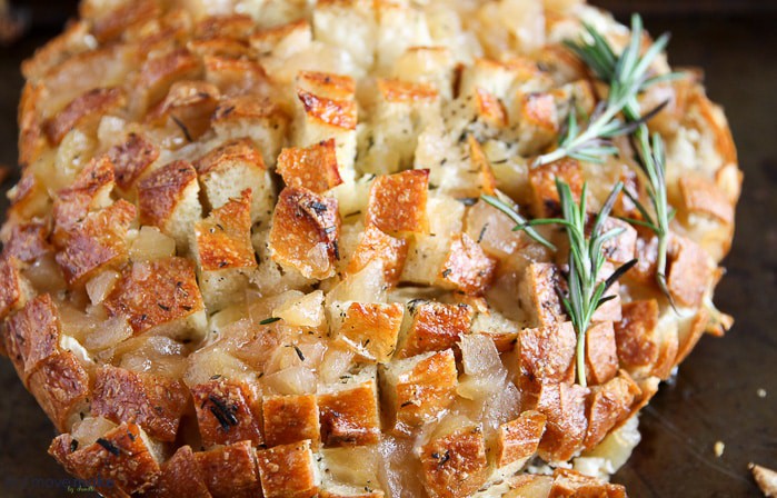 A close-up of a baked dish featuring a round loaf of bread scored in a criss-cross pattern, filled with melted cheese, and garnished with sprigs of fresh rosemary. The bread has a golden, crispy crust.