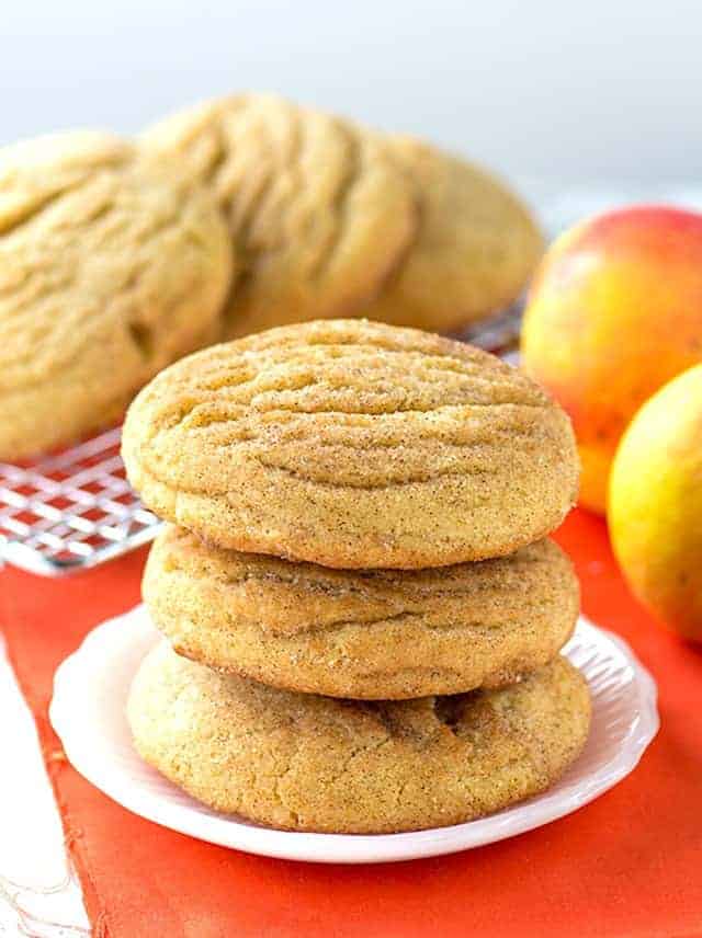 A stack of three golden snickerdoodle cookies on a white plate, set against an orange cloth. Behind the stack, more cookies rest on a cooling rack, with two apples beside them.