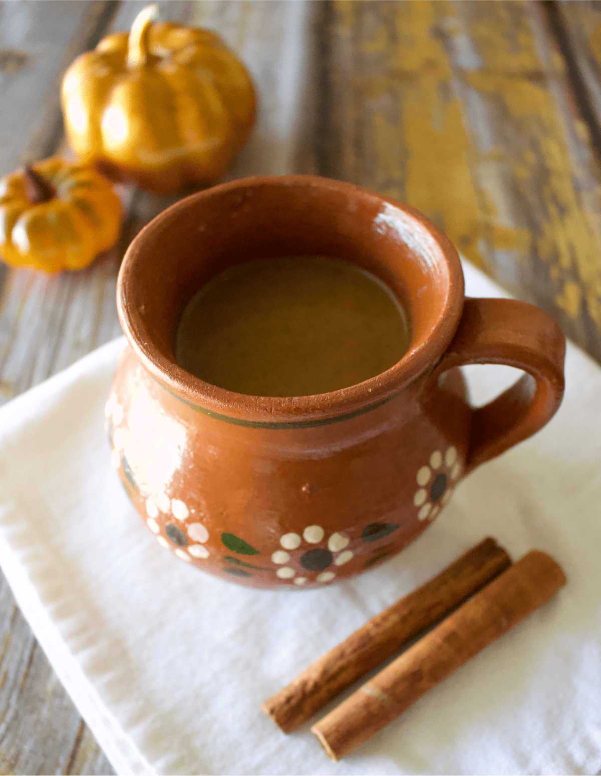 A brown ceramic mug with floral patterns filled with pumpkin spice latte sits on a white cloth. Two cinnamon sticks are placed beside it, and small decorative pumpkins, reminiscent of pumpkin puree dessert recipes, adorn the rustic wooden surface in the background.