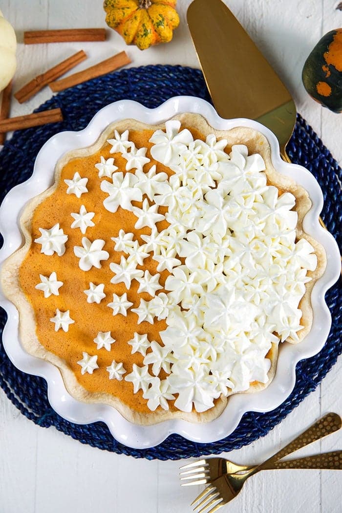 A pie crowned with a gradient of whipped cream stars, transitioning from sparse to dense, showcases an artful touch. It rests on a blue woven mat surrounded by cinnamon sticks, small pumpkins, and gold utensils on a white wooden table—a perfect addition to any pumpkin puree dessert repertoire.