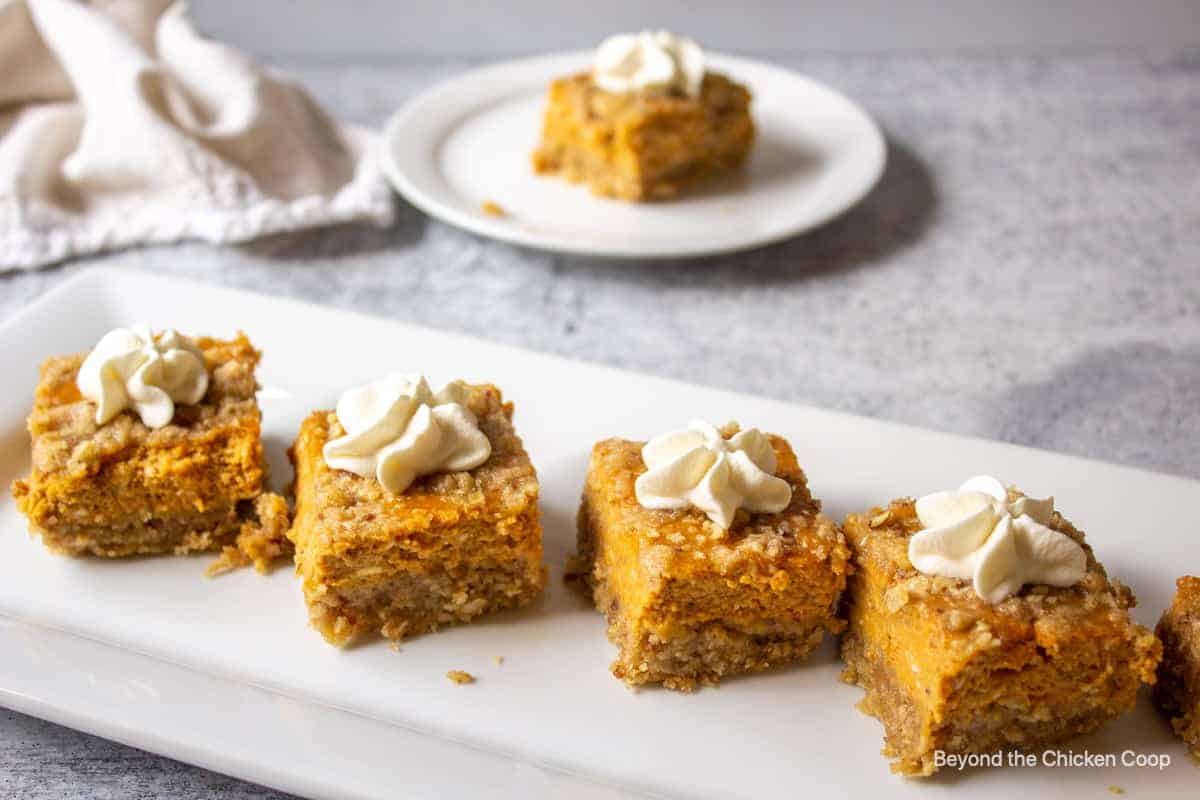 A plate of pumpkin bars made with rich pumpkin puree, topped with dollops of whipped cream. In the background, a white plate holds another piece of the dessert, placed on a gray surface. A crumpled beige napkin is nearby—a tempting treat for those seeking delicious dessert recipes.