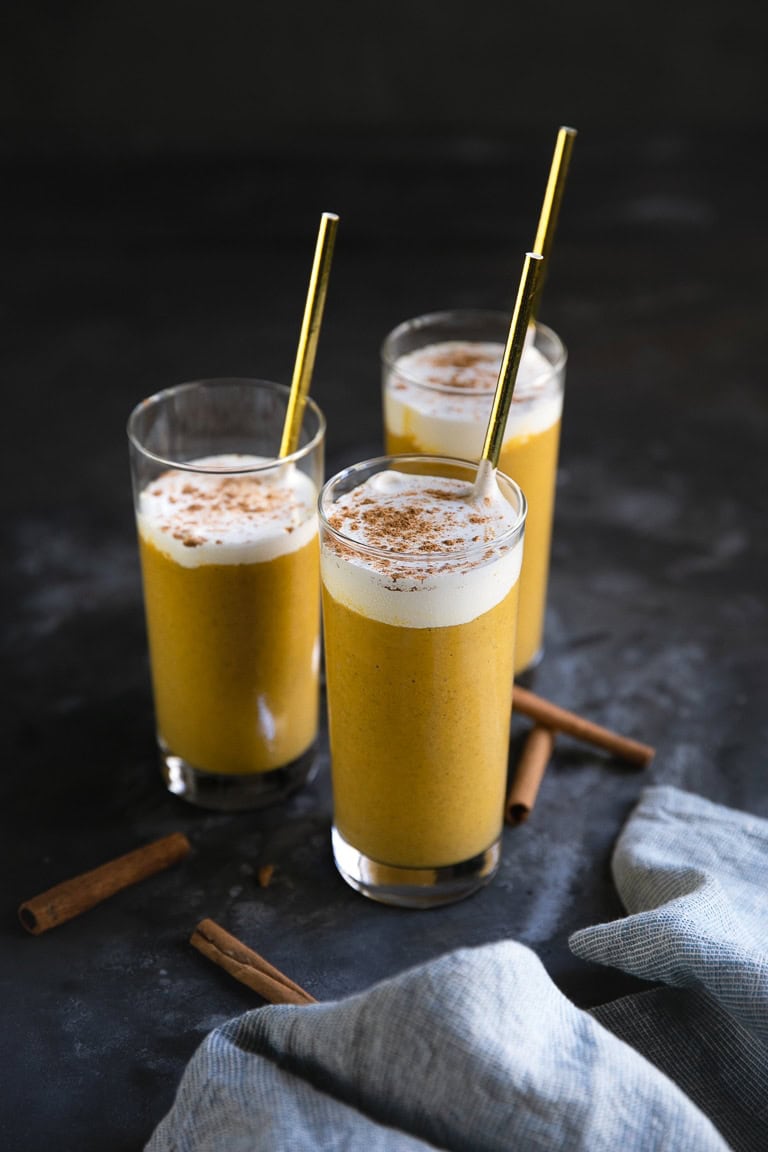 Three tall glasses of a frothy pumpkin spice drink, reminiscent of pumpkin puree dessert recipes, sit on a dark surface. Each is topped with a sprinkle of cinnamon and served with a metal straw. Cinnamon sticks and a striped cloth are casually placed nearby, accentuating the cozy scene.