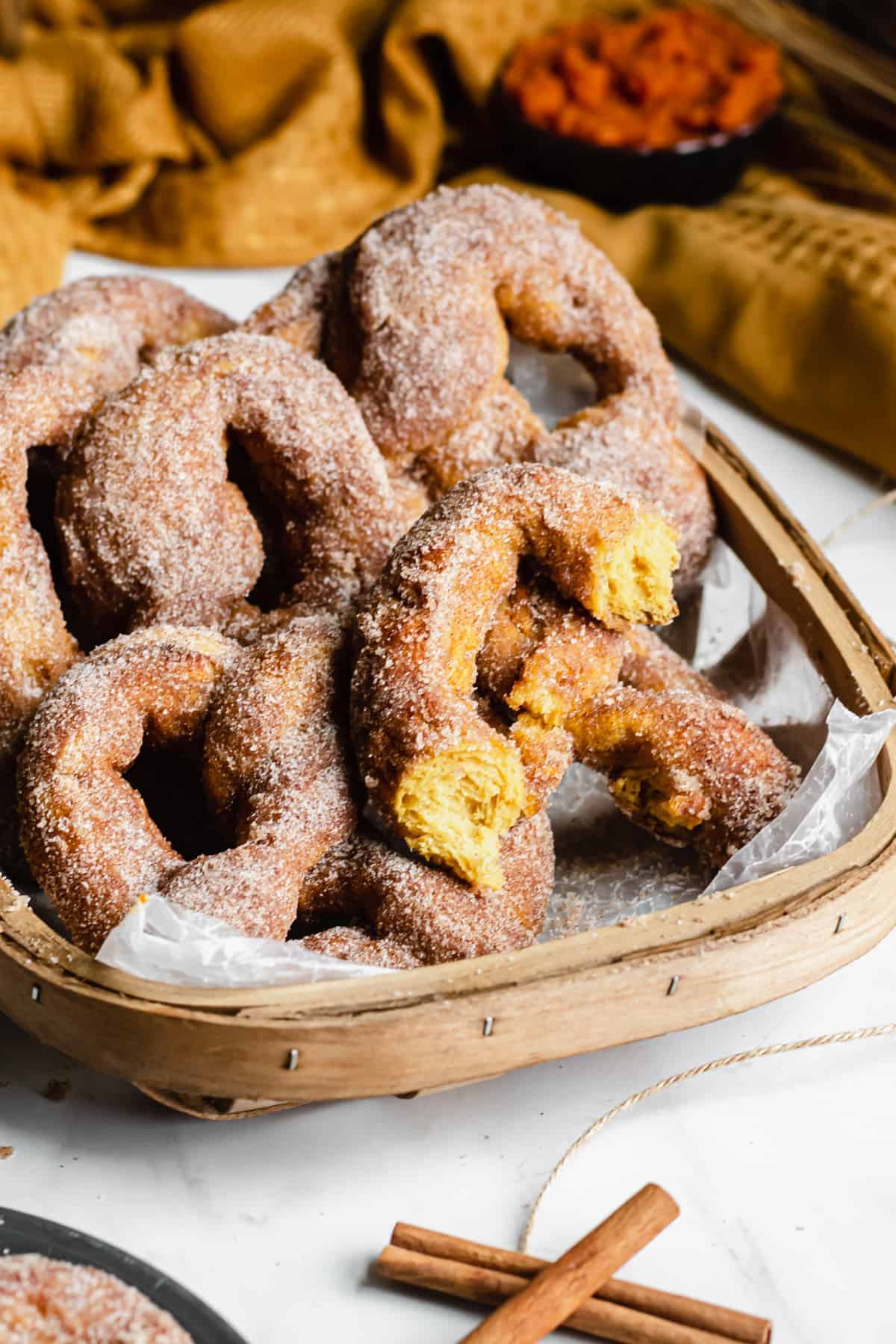 A basket filled with sugar-coated pumpkin donuts, some with bites taken out, is placed on a wooden surface. A few cinnamon sticks lie nearby, reminiscent of cozy pumpkin puree dessert recipes. A cloth napkin is partially visible in the background.