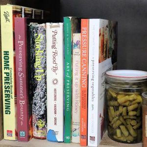 A variety of books about canning on a shelf with a jar of green beans.