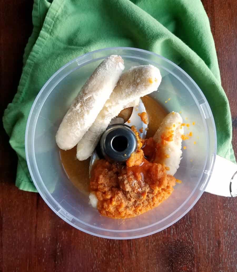 A food processor bowl containing frozen bananas, pumpkin puree, and a dusting of spices sits on a wooden table with a green cloth beneath it, inspiring delightful pumpkin puree dessert recipes.