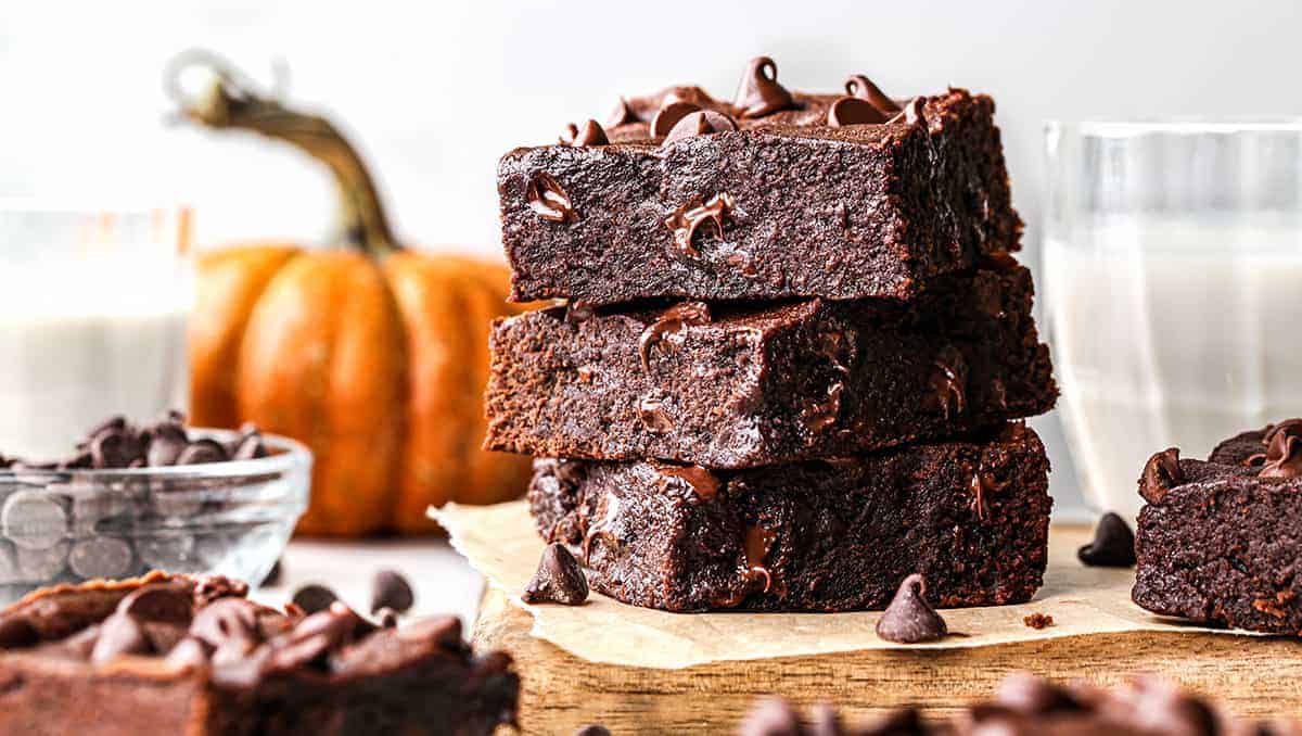 Stack of chocolate brownies with chocolate chips on top, nestled on parchment paper. A small pumpkin and a glass of milk accompany the scene, hinting at cozy pumpkin puree dessert recipes perfect for autumn. A bowl of chocolate chips adds to the indulgent setting.