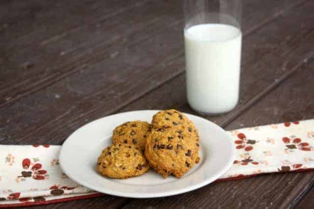 A plate of three chocolate chip cookies, with a hint of pumpkin puree, sits on a floral napkin atop a wooden table. A glass of milk is in the background.