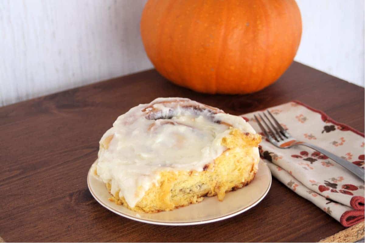 A cinnamon roll topped with creamy icing sits on a plate on a wooden table. Nearby, a pumpkin and a folded napkin with a fork evoke the essence of autumn. This cozy pumpkin puree dessert recipe brings warmth to any fall gathering.