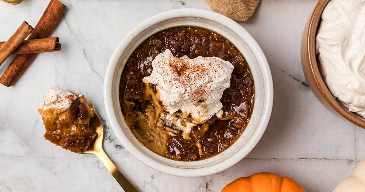 A bowl of pumpkin bread pudding, enhanced with pumpkin puree for extra flavor, is topped with whipped cream and cinnamon on a marble surface. A spoon with a portion of this delightful dessert lies beside the bowl, while a small pumpkin and cinnamon sticks add seasonal charm nearby.