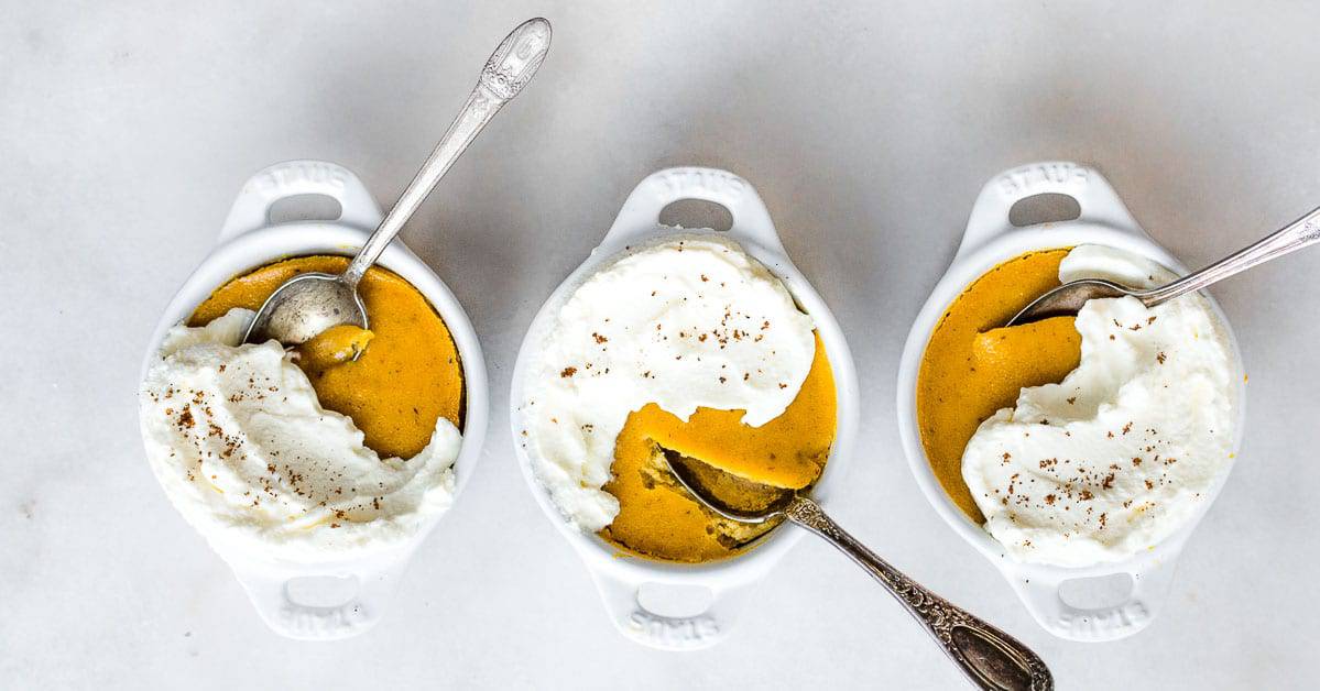 Three small white dishes filled with pumpkin pie filling are topped with whipped cream and scattered with a pinch of spices. Each dish, crafted from delightful pumpkin puree, has a silver spoon resting on the creamy topping. The dishes are neatly arranged on a light surface—perfect for dessert recipe inspiration.