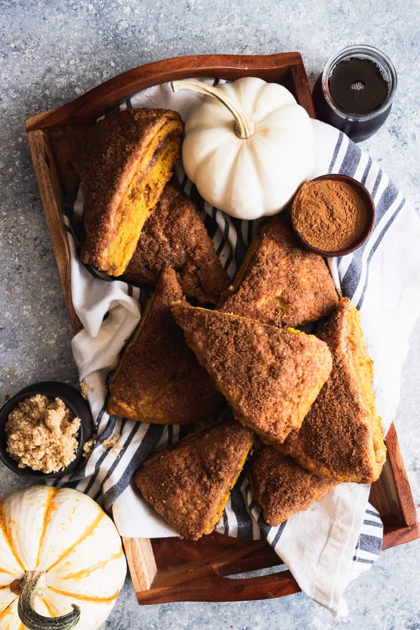 A wooden tray presents pumpkin scones made with creamy pumpkin puree, surrounded by petite white pumpkins, a bowl of cinnamon, brown sugar, and a dark beverage in a glass. A striped cloth lines the tray, enhancing the warm ambiance typical of autumn dessert recipes.