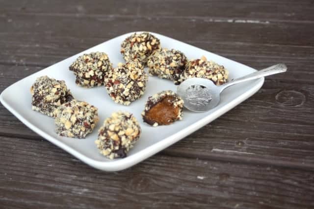 A white square plate presents nine chocolate truffles, each glistening with chopped nuts. A small silver spoon rests elegantly beside them, hinting at the artistry akin to pumpkin puree dessert recipes. The background is a contrasting dark wooden surface.