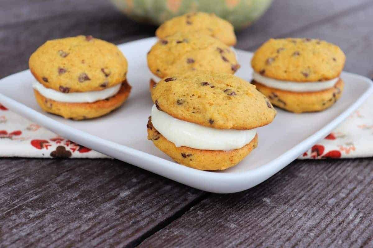 A white plate with four pumpkin whoopie pies, crafted from pumpkin puree dessert recipes, sits on a wooden table. The whoopie pies have chocolate chips and a soft texture, while a floral napkin is partially visible underneath the plate.