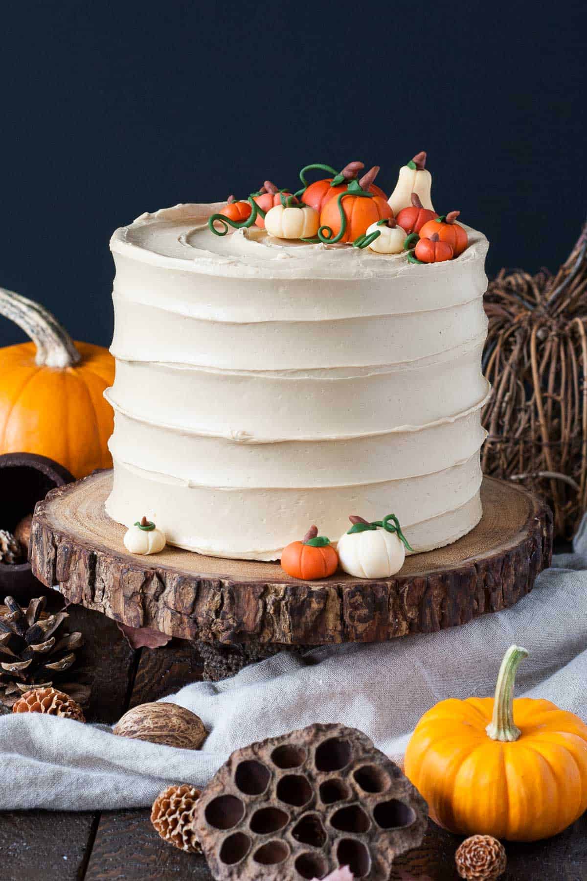 A rustic cake, highlighting pumpkin puree dessert recipes, features smooth beige frosting and is adorned with fondant pumpkins. Resting on a wooden slab, it’s surrounded by autumn-themed items like pumpkins, pine cones, and dried lotus pods against a dark background.