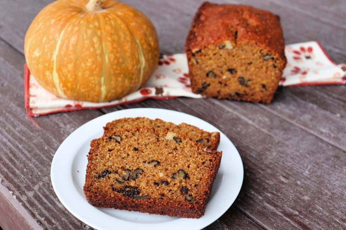 A plate with two slices of pumpkin bread, showcasing visible nuts, rests on a wooden table. In the background, a whole pumpkin and a loaf sit on a patterned cloth, evoking thoughts of delightful pumpkin puree dessert recipes.