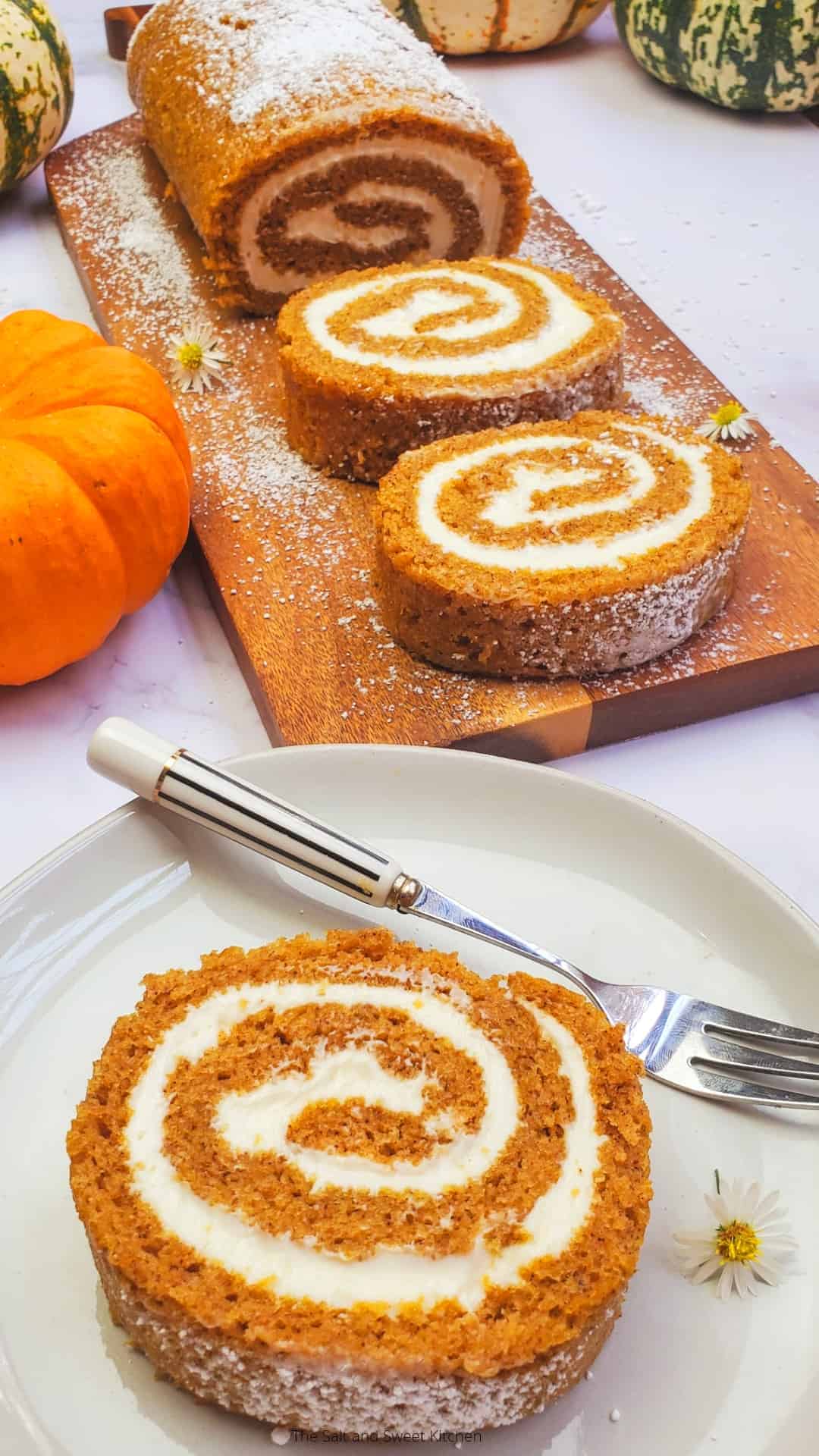 A pumpkin roll cake made with luscious pumpkin puree is sliced and displayed on a wooden board. The pieces are elegantly arranged on a white plate, fork at the ready. Decorative pumpkins and daisies complete the festive autumn setting, offering inspiration for your dessert recipe collection.