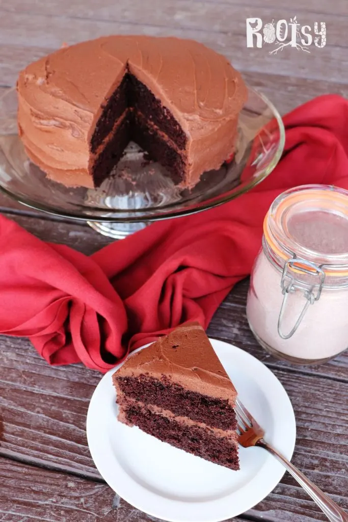 A slice of chocolate cake with chocolate frosting sits on a plate with a fork. In the background the remaining cake sits on a cake plate with a jar of chocolate cake mix. 