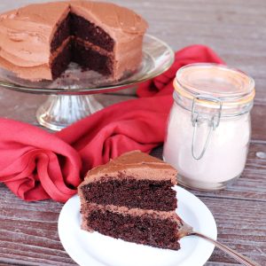 A slice of chocolate cake with chocolate frosting on a plate with a fork. In the background the remaining cake sits on an cake plate next to a jar of chocolate cake mix.