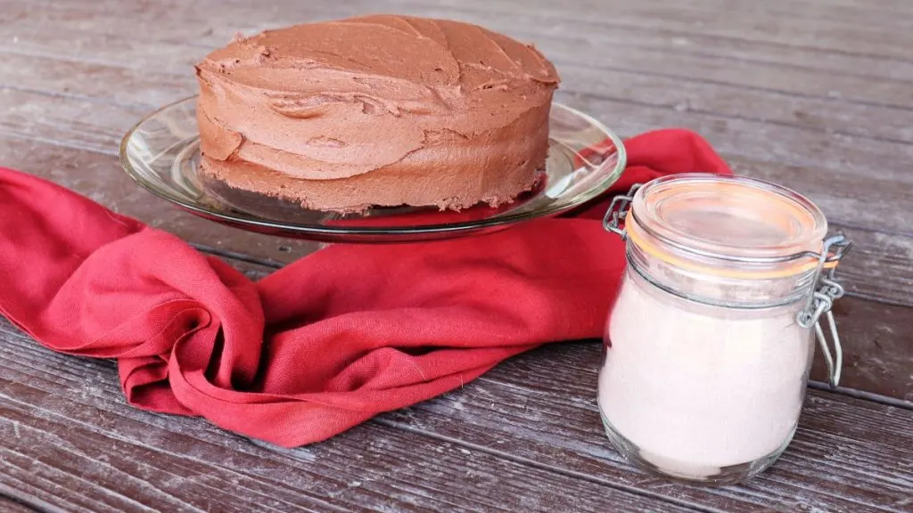 A frosted chocolate cake sits on a glass cake plate with a red cloth surrounding it. A jar of cake mix sits to the right. 